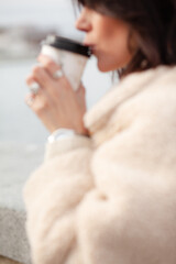 A beautiful girl with a stylish bob haircut is walking around the city, drinking coffee or tea, sitting against the sea. The woman is wearing a black leather, sweater, torn jeans and boots