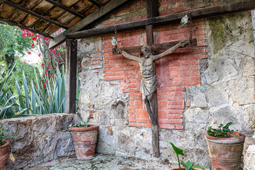 Huge old wooden cross with crucified Christ in shed of ranch, tile roof, worn brick walls, clay pots with small green plants, red bougainvillea and trees in background, sunny day in Jalisco Mexico