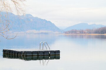 Spokój nad górskim jeziorem zimą. Drewniana platforma ze srebrną drabinką dla pływaków i plażowiczów. W tle zamglone góry. - obrazy, fototapety, plakaty