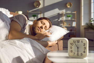 Guy is sleeping sweetly, lying in his cozy bed at home in the living room. Attractive bearded young man is lying in bed, his head on pillow and covered with warm blanket. Sunny early morning, at 6 am.