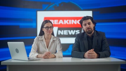Male and female presenters in the studio. Man and woman news hosts sit at desk presenting daily events, reporting on air, breaking news background at the back.