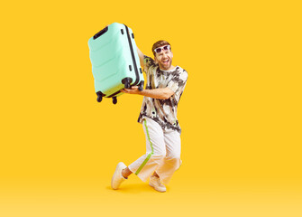 Full body photo of a funny young happy man in sunglasses carrying his blue suitcase is going on summer holiday trip and having fun on a yellow background. Vacation and travel concept. - obrazy, fototapety, plakaty