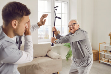 Angry elderly senior man going to hit young male caregiver or nurse with his crutch. Enraged...