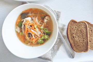 View of a healthy soup of vegetables and meat in a white plate on the table with bread and spoon with copy space. Traditional soup with broccoli, carrots, pieces of meat, served in plate. 