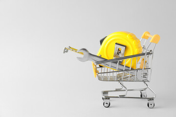 Metal wrench and measuring tape in shopping cart on light grey background, space for text
