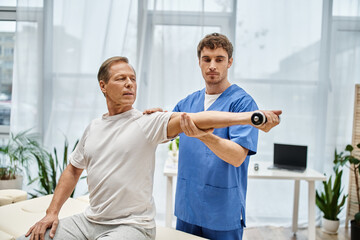Fototapeta premium handsome doctor in blue robe helping his mature patient in casual attire to use dumbbell in hospital