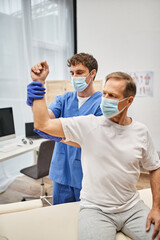 hardworking doctor with mask and gloves helping mature patient to rehabilitate his muscles in ward