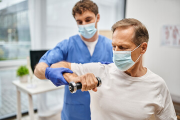 Fototapeta na wymiar devoted handsome doctor with mask and gloves helping his patient to use dumbbells on appointment