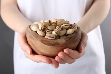 Woman holding tasty pistachios in bowl on grey background, closeup