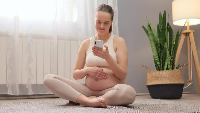 Cheerful pregnant woman holding mobile phone touching belly while scrolling online holding smartphone chatting with friends or choosing something in web store