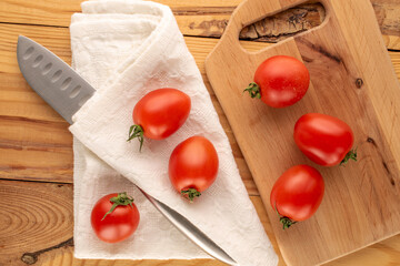 Several cocktail tomatoes with a knife on a white napkin and a wooden kitchen board on a wooden table, macro, top view.