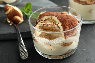 Delicious tiramisu in glass and spoon on black table, closeup