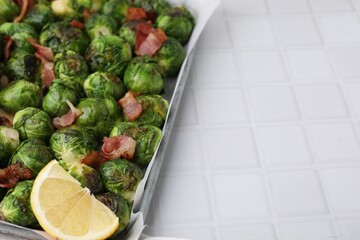 Delicious roasted Brussels sprouts, bacon and lemon in baking dish on white tiled table, closeup. Space for text