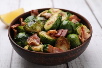 Delicious roasted Brussels sprouts and bacon in bowl on light wooden table, closeup