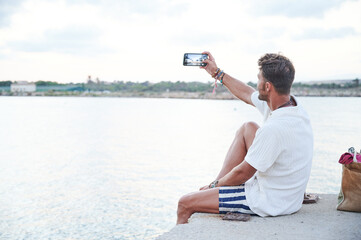 Good looking 40 year old man dressed in summer clothes uses his smartphone to take a sunset photo on a seaside breakwater during his vacation. - obrazy, fototapety, plakaty