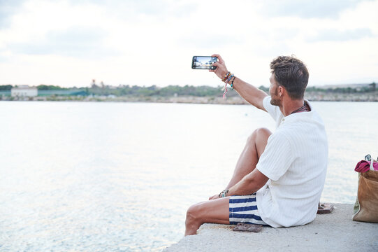 Good looking 40 year old man dressed in summer clothes uses his smartphone to take a sunset photo on a seaside breakwater during his vacation.