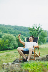 Cute smiling man on folding wooden chair sitting in nature and resting. The concept of recreation...