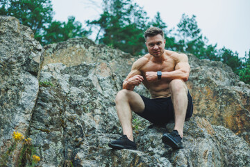 A muscular young man sits on the rocks near the forest. Relief and muscular physique of a man.