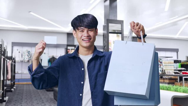 An Asian Shopping Man With A White Card Holding Shopping Bags Up And Smiling To Camera While Standing In Clothing Store