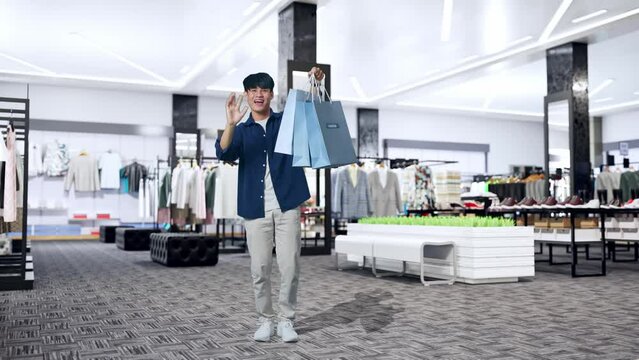 Full View Of An Asian Shopping Man Holding Shopping Bags Up, Waving Hands And Smiling To Camera While Standing In Clothing Store