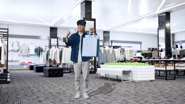 Full View Of An Asian Shopping Man With A White Card Holding Shopping Bags Up And Smiling To Camera While Standing In Clothing Store