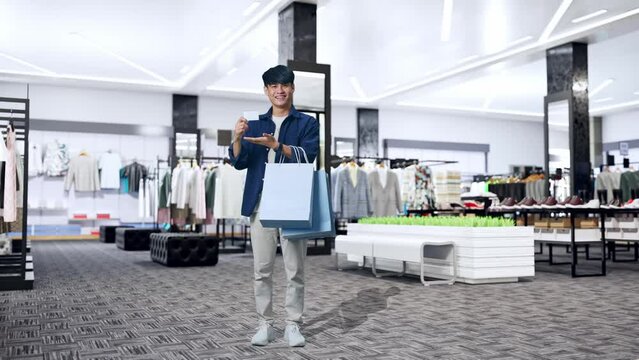 Full View Of An Asian Shopping Man With A White Card And Shopping Bags Smile To Camera While Standing In Clothing Store