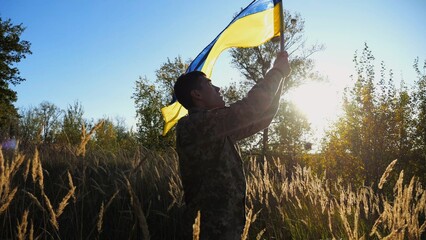 Male military in uniform waving flag of Ukraine at countryside. Young soldier of ukrainian army lifting blue-yellow banner as symbol of victory against russian aggression. Invasion resistance concept. - 759083910