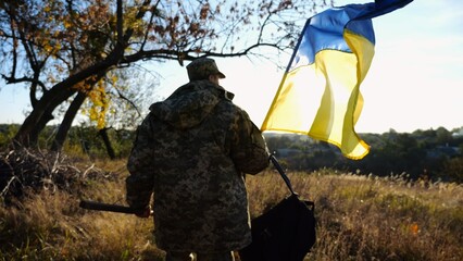Young male military in uniform walks with waving flag of Ukraine at countryside. Soldier of ukrainian army going with blue-yellow banner outdoor. Concept of victory against russian aggression. - 759083984