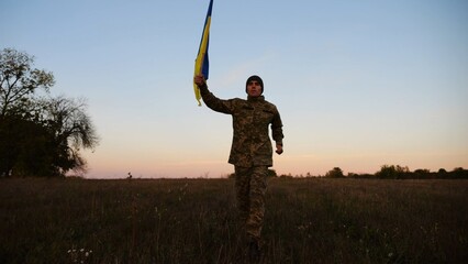 Soldier of ukrainian army running with raised blue-yellow banner on field at dusk. Young male military in uniform jogging with flag of Ukraine at meadow. Victory against russian aggression concept - 759084360