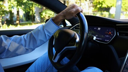 Hands of businessman holds steering wheel while driving electric car at city. Male businessperson operating electrical vehicle on urban road. Young man commuting to work at sunny day. Close up - 759085359