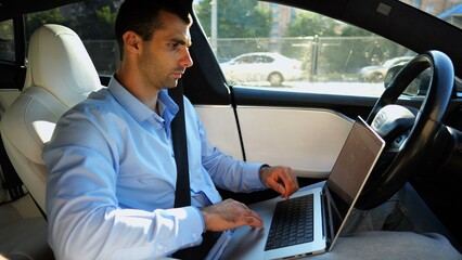 Handsome entrepreneur working on notebook while riding an autonomous self driving electric car at urban road. Male businessman typing text on laptop during riding on electrical vehicle with autopilot - 759085989