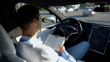 Male businessperson reading book during riding on electrical vehicle with autopilot at urban road. Successful businessman improving his knowledge while riding an autonomous self driving electric car - 759086322