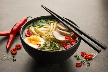 Delicious ramen with meat in bowl, chopsticks and chili peppers on grey textured table. Noodle soup