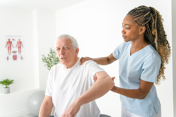 Obraz premium Black physiotherapist helping senior man with in clinic. Elderly man undergoing physiotherapy treatment for injury