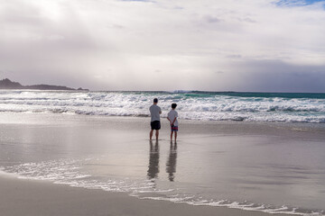 Man and boy standing on a beach watching breaking waves.
