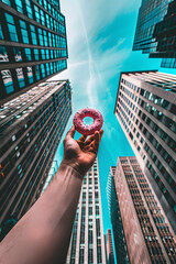 A hand holds a donut towards the skyscrapers in the city. Sunny blue sky.