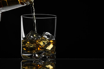 Pouring whiskey into glass with ice cubes at table against black background, space for text