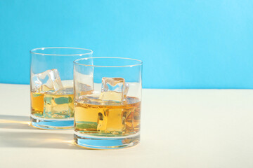 Whiskey with ice cubes in glasses on white table against light blue background, space for text