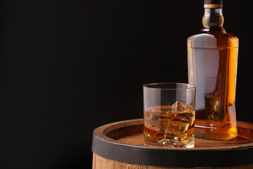 Whiskey with ice cubes in glass and bottle on wooden barrel against black background, space for text
