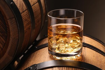 Whiskey with ice cubes in glass on wooden barrel against dark background, closeup