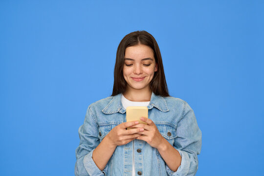 Happy pretty gen z Latin teen girl, smiling teenage student with brunette hair wearing denim jacket holding smartphone, using cellphone looking at mobile phone standing isolated on blue background.