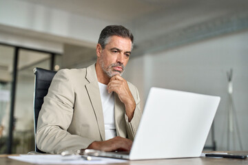 Busy mature professional business man ceo looking at laptop working at desk. Serious middle aged executive manager investor using computer thinking on digital financial investing risks in office.