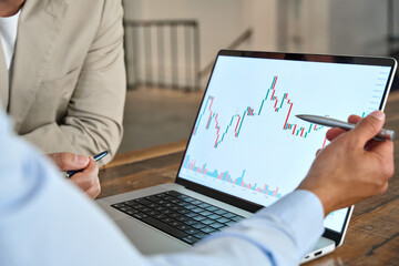 Financial advisor explaining invest stock market data consulting investor. Two business men analysts doing finance trading analysis pointing at chart on laptop screen working in office. Close up.
