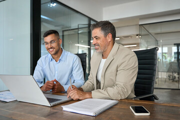 Two busy happy business men partners working together talking using laptop in office. Middle aged Latin male manager and young worker looking at computer discussing business project at office meeting.