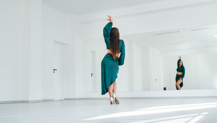 Graceful woman dressed in dark green Latin dancing dress doing elegant dance pas in white color big hall with big mirror wall. People's expressions during dancing, beauty of woman's body concept image