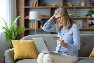 A mature woman looking surprised and anxious while holding a credit card and using a laptop on her...