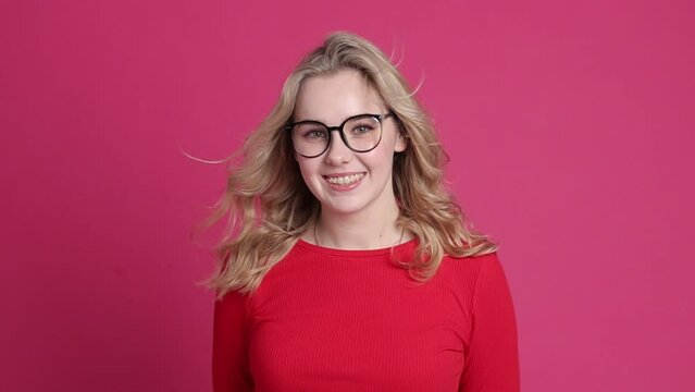 Cheerful young caucasian woman with transparent ceramic braces on teeth isolated on pink backdrop. Blond girl in eyeglasses looks at camera. Windy effect with fan. Beauty, health and fashion concept.