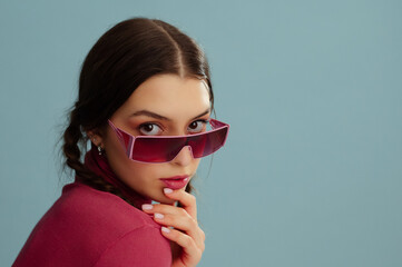 Fashionable confident woman wearing trendy pink wraparound 90s style sunglasses, turtleneck, posing on blue background. Studio fashion portrait. Copy, empty, blank space for text
