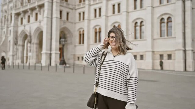 A stylish young woman wearing glasses poses with a playful smile against the backdrop of the historic budapest parliament in hungary.