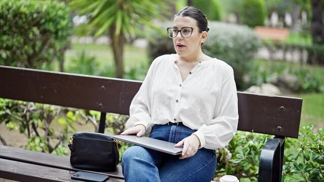 Young beautiful hispanic woman using laptop sitting on a bench finishing work at the park